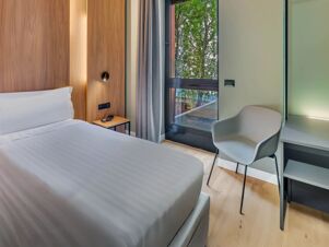 White bed with wooden headboard, bedside table with phone, gray chair, window with green trees view in B&B Hotel Milano Portello
