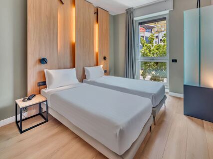 Two single beds with white linens and pillows, wooden headboard with wall lamps, window and small bedside table in B&B Hotel Milano Portello room