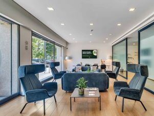Modern lounge area at B&B Hotel Milano Portello with blue upholstered armchairs, sofas, a coffee table, and large windows showing outside trees and cars