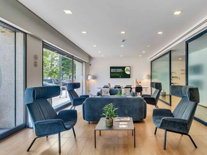 Modern lounge area at B&B Hotel Milano Portello with blue upholstered armchairs, sofas, a coffee table, and large windows showing outside trees and cars