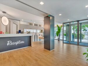 Reception area with wood flooring, white and gray reception desk, round hanging light, potted plants, glass doors at B&B Hotel Milano Portello