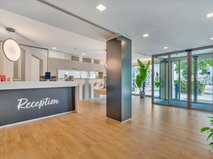 Reception area with wood flooring, white and gray reception desk, round hanging light, potted plants, glass doors at B&B Hotel Milano Portello