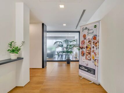 Bright hallway with wooden floor and potted plants next to a B&B Hotel Milano Portello breakfast advertisement banner