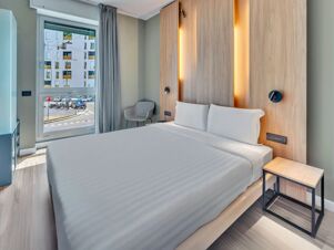 Modern hotel room at B&B Hotel Milano Portello with a neatly made bed, wooden headboard with lights, grey chair, and window showing outside view