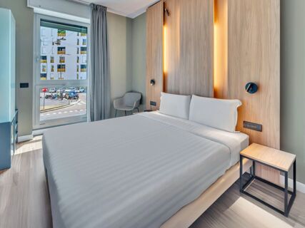 Modern hotel room at B&B Hotel Milano Portello with a neatly made bed, wooden headboard with lights, grey chair, and window showing outside view