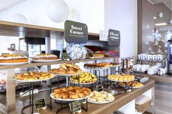 Various baked goods including croissants, muffins, and cakes displayed on wooden shelves in B&B Hotel Milano Portello