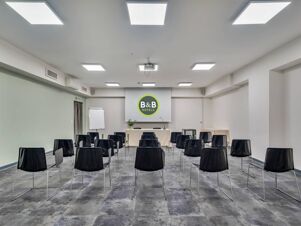 Conference room setup with black chairs, a projector screen displaying B&B Hotels logo, a white table, and flip chart at B&B Hotel Milano Portello