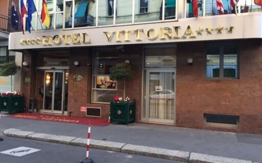 Hotel Vittoria entrance with gold signage, flags above, glass doors, and planters with flowers on the sidewalk