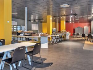 Dining area with white tables and black chairs, yellow pillars, food display counters, and kitchen equipment at B&B Hotel Milano Ornato