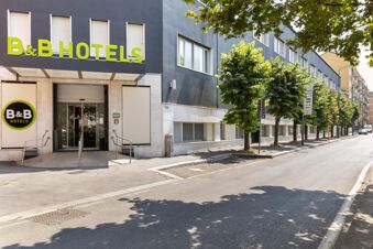 Entrance of B&B Hotel Milano Ornato with large green signage, glass doors, gray building facade, and a tree-lined street alongside