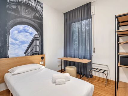 Simple hotel room at B&B Hotel Milano Ornato with single bed, wall art, desk, stool, and a window with grey curtains