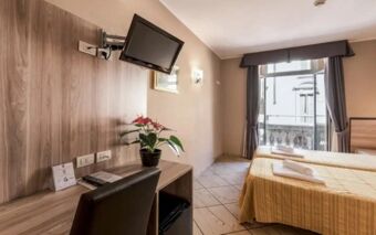 Hotel Brianza room with twin beds, yellow bedspreads, a desk with a chair, a wall-mounted TV, and a balcony window