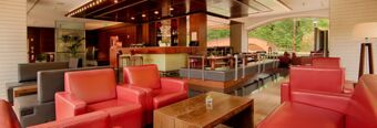 Lounge area inside NH Milano 2 with red and gray armchairs, wooden tables, a bar, and large windows showing greenery outside