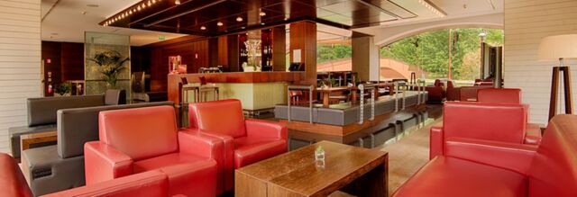 Lounge area inside NH Milano 2 with red and gray armchairs, wooden tables, a bar, and large windows showing greenery outside