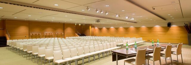 A large conference room at NH Milano 2 with rows of beige chairs and a table with water bottles and papers