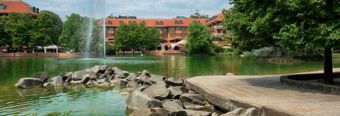 Water fountain in a pond with rocks, trees, and red-brick buildings including NH Milano 2 in the background under a blue sky