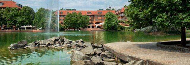 Water fountain in a pond with rocks, trees, and red-brick buildings including NH Milano 2 in the background under a blue sky