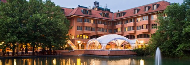 Hotel NH Milano 2 with peach-colored walls, red roof, large trees, white canopy, and a water fountain in front at dusk