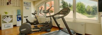 Small fitness room with treadmill, elliptical, exercise bike, large windows, water dispenser, towels, and a wellness poster at NH Milano 2