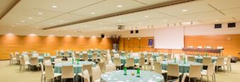 Large conference room at NH Milano 2 with round tables, beige chairs, green water bottles, and a projection screen on wood-paneled walls