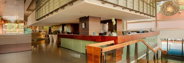 Spacious reception area of NH Milano 2 with a large wooden front desk, glass railing, potted plants, and modern lighting fixtures
