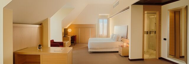 Spacious hotel room at NH Milano 2 with a large bed, wooden desk, armchair, a desk lamp, and an open bathroom door