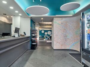 Reception area with a gray counter, map on the wall, product display shelves, blue chairs, and a refrigerator at B&B Hotel Torino President