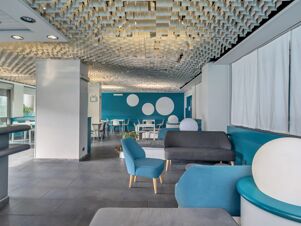 Spacious seating area with blue and gray chairs and sofas, white tables, and geometric ceiling at B&B Hotel Torino President