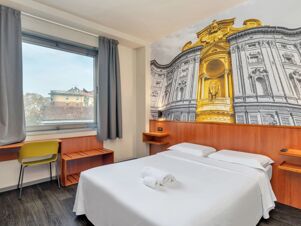 Hotel room at B&B Hotel Torino President with a double bed, white linens, rolled towels, desk, yellow chair, window, and large architectural wall mural