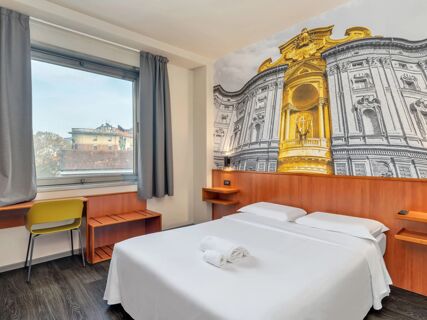 Hotel room at B&B Hotel Torino President with a double bed, white linens, rolled towels, desk, yellow chair, window, and large architectural wall mural