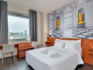 Room interior at B&B Hotel Torino President showing a white double bed, towel rolls, a desk with chair, window with gray curtains, and wall art with statues