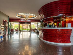 Spacious hotel lobby with glossy red curved reception desk, chandelier, glass entrance, seating area, and snack shelves at B&B Hotel Borgaro Torinese