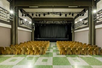 Conference room with rows of yellow covered chairs, green tiled floor, stage with black curtains and spotlights at B&B Hotel Borgaro Torinese
