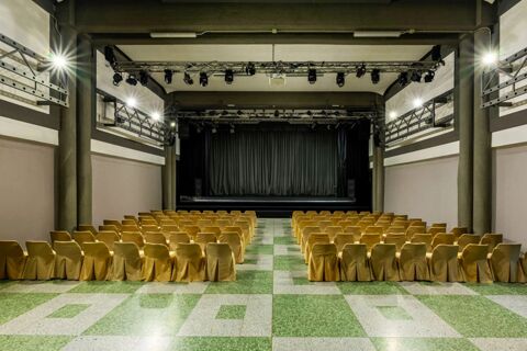 Conference room with rows of yellow covered chairs, green tiled floor, stage with black curtains and spotlights at B&B Hotel Borgaro Torinese