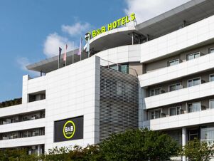 Modern white multi-story building with balconies, trees in front, flags, and B&B Hotel Borgaro Torinese signs on top and side