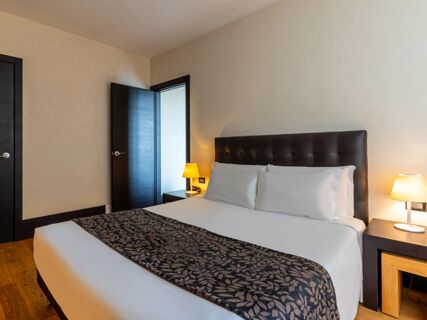 Bed with white linens and a dark patterned runner beside a nightstand with a lamp and an open door in B&B Hotel Borgaro Torinese