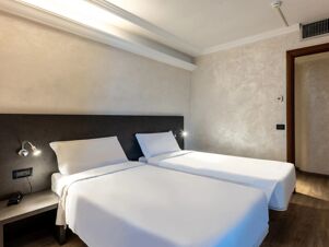 Two single beds with white linens and pillows in a minimalistic room at B&B Hotel Borgaro Torinese with bedside lamps and air vent