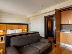 Modern hotel room at B&B Hotel Borgaro Torinese with a bed, wooden headboard, bedside lamps, sofa, and kitchenette area