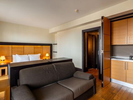 Modern hotel room at B&B Hotel Borgaro Torinese with a bed, wooden headboard, bedside lamps, sofa, and kitchenette area