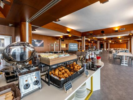 Buffet area with croissants, packaged bread, beverages, toaster, and seating in a spacious room at B&B Hotel Borgaro Torinese