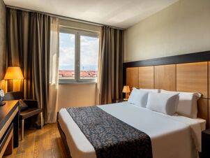 Double bed with white linens and patterned runner near window with curtains and lamps in B&B Hotel Borgaro Torinese room