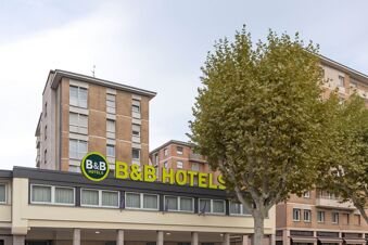 B&B Hotel Como City Center building with large yellow and green signage on the roof and a tree in front under a cloudy sky