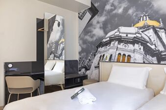 White bedding with folded towel and remote on bed, beige chair, black desk, and large wall photo in B&B Hotel Como City Center