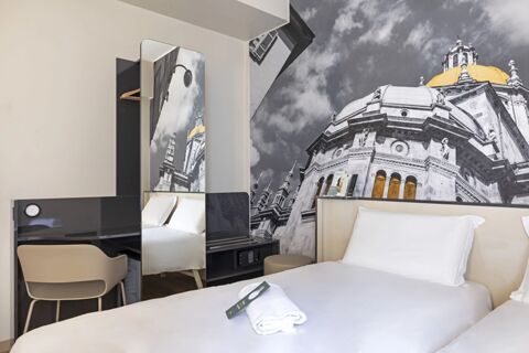 White bedding with folded towel and remote on bed, beige chair, black desk, and large wall photo in B&B Hotel Como City Center
