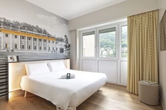 Double bed in a modern room with wall art, patterned curtains, a window view, and a "Please tidy my room" card at B&B Hotel Como City Center