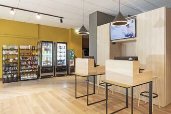 Reception area of B&B Hotel Como City Center with wooden desks, hanging lights, snack racks, and two refrigerators visible on a yellow wall