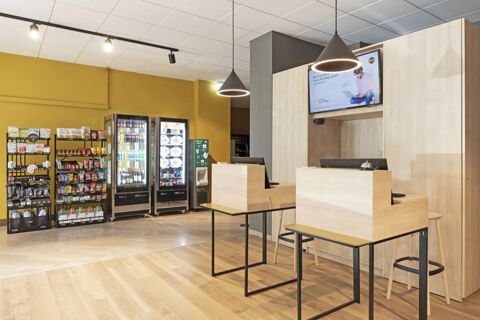 Reception area of B&B Hotel Como City Center with wooden desks, hanging lights, snack racks, and two refrigerators visible on a yellow wall