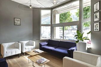 A seating area inside B&B Hotel Como City with white armchairs, a dark purple sofa, wooden tables, large windows, and framed art on the walls