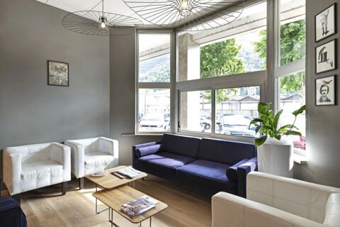 A seating area inside B&B Hotel Como City with white armchairs, a dark purple sofa, wooden tables, large windows, and framed art on the walls