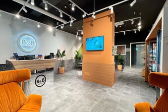 Reception desk with computers, a glowing B&B Hotels sign, plants in pots, orange chairs, and a mounted screen at B&B Hotel Como Baradello